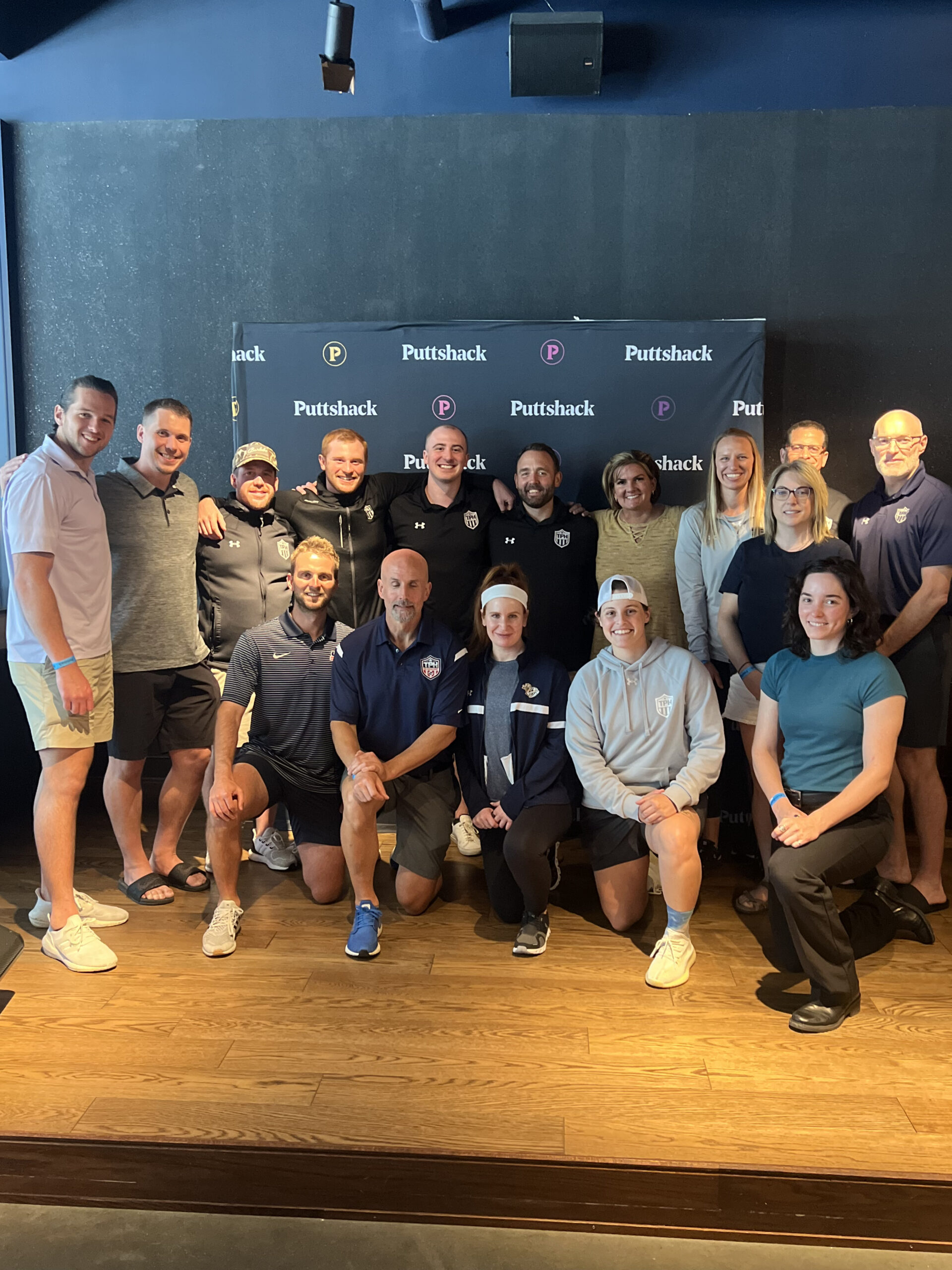 A group of about 20 people, casually dressed, pose together and smile in front of a Puttschack-branded backdrop on a wooden floor, representing TPH Academy in an indoor setting.