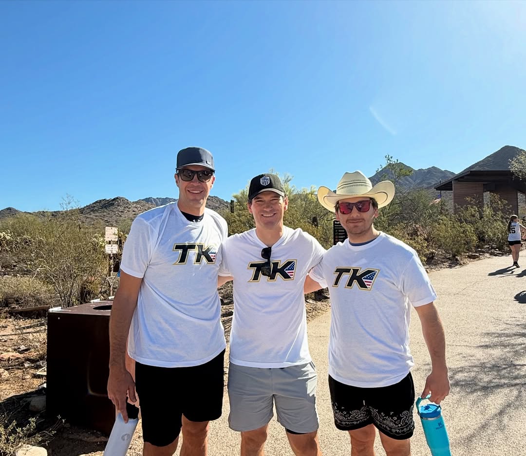 Three men stand outdoors on a sunny day, posing and smiling for a photo in their white TPH Academy shirts and shorts; one wears a cowboy hat, another a baseball cap, and the third sunglasses. Desert mountains stretch out in the background.