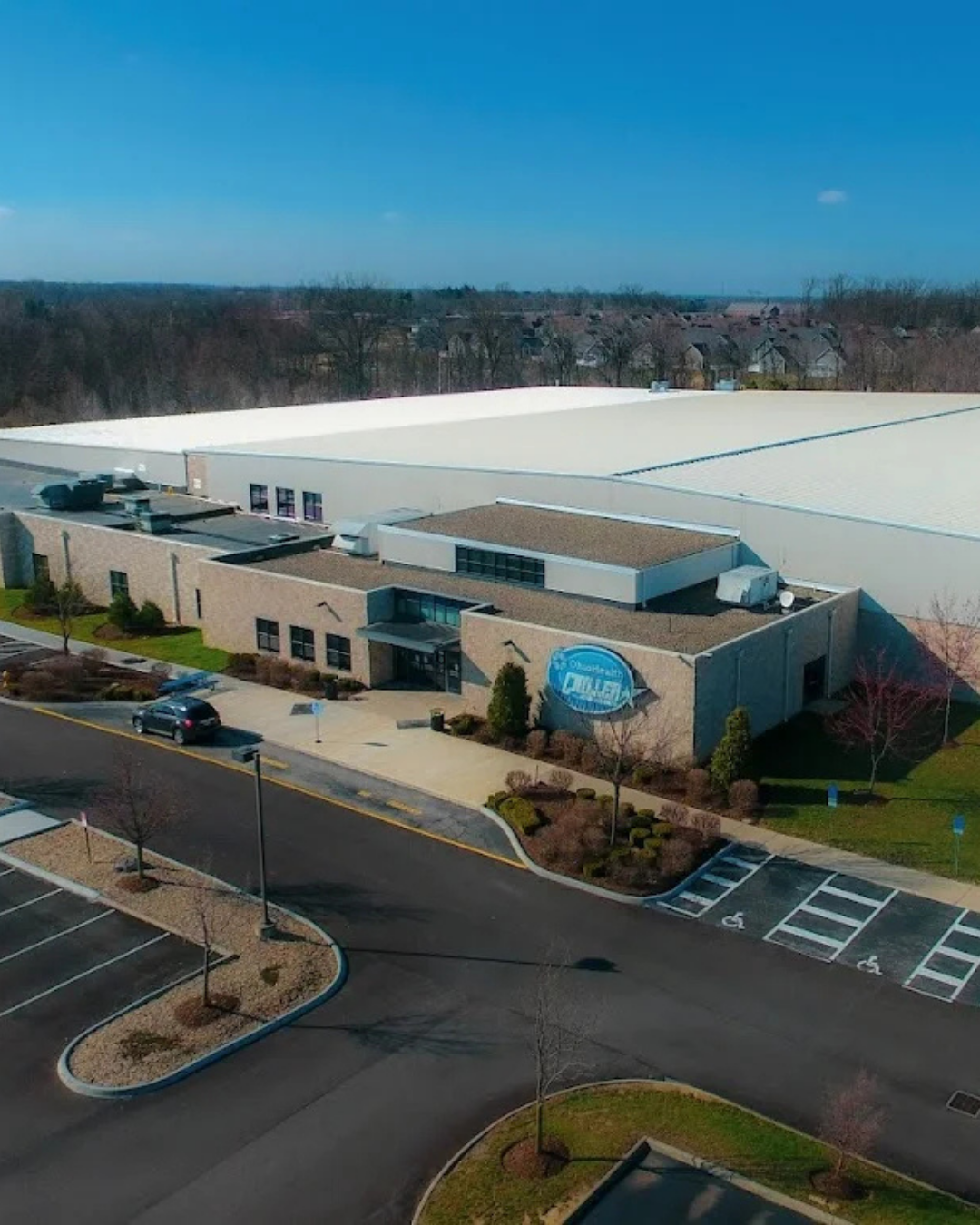 Aerial view of a modern building with a sign reading Chiller, home to TPH Academy, surrounded by a parking lot with several cars and accessible spaces, with trees and houses in the background under a clear blue sky.