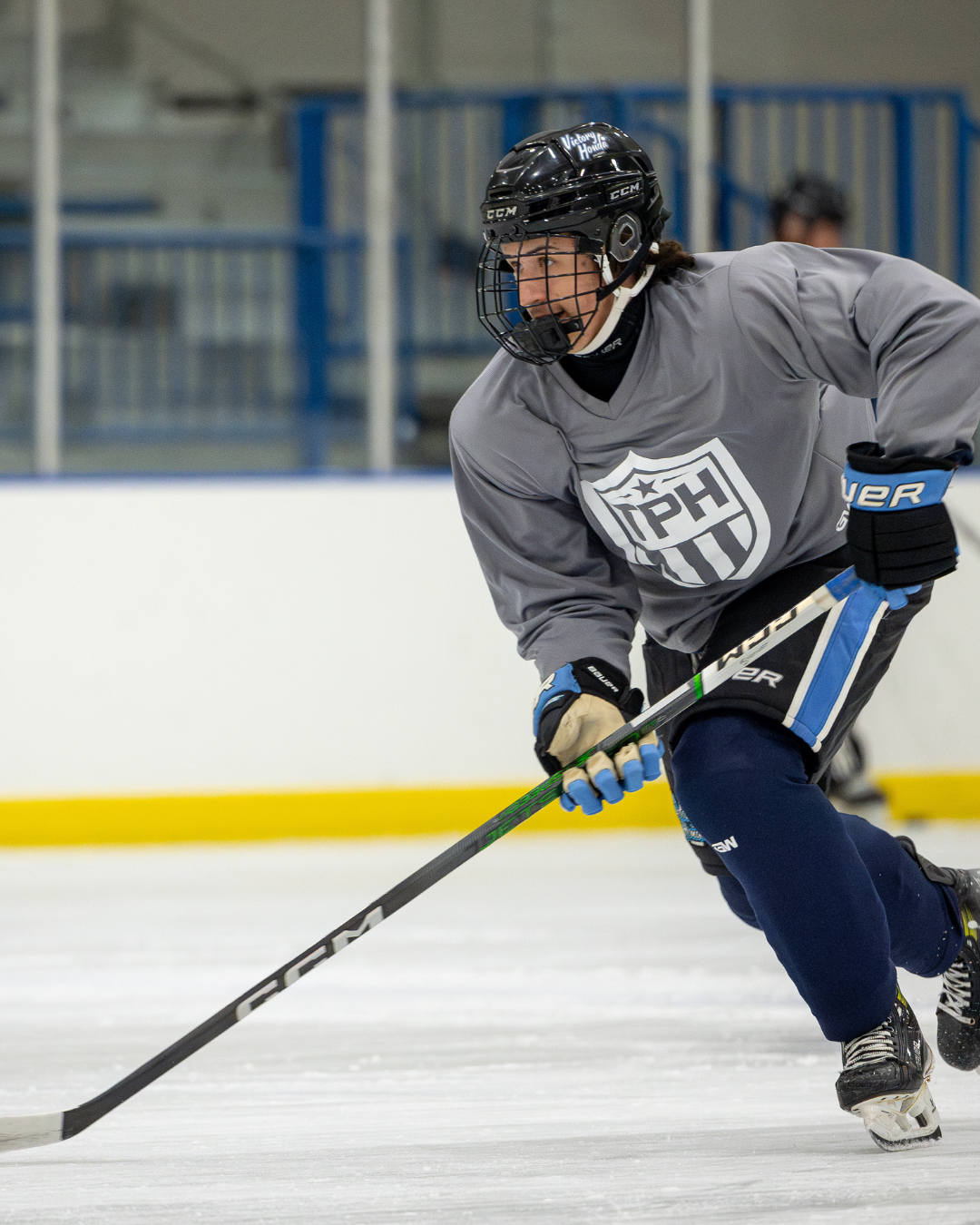 A hockey player in a gray jersey, black helmet, and blue gloves skates on the ice holding a hockey stick. The background shows part of the TPH Academy rink with blue railings and another player slightly out of focus.