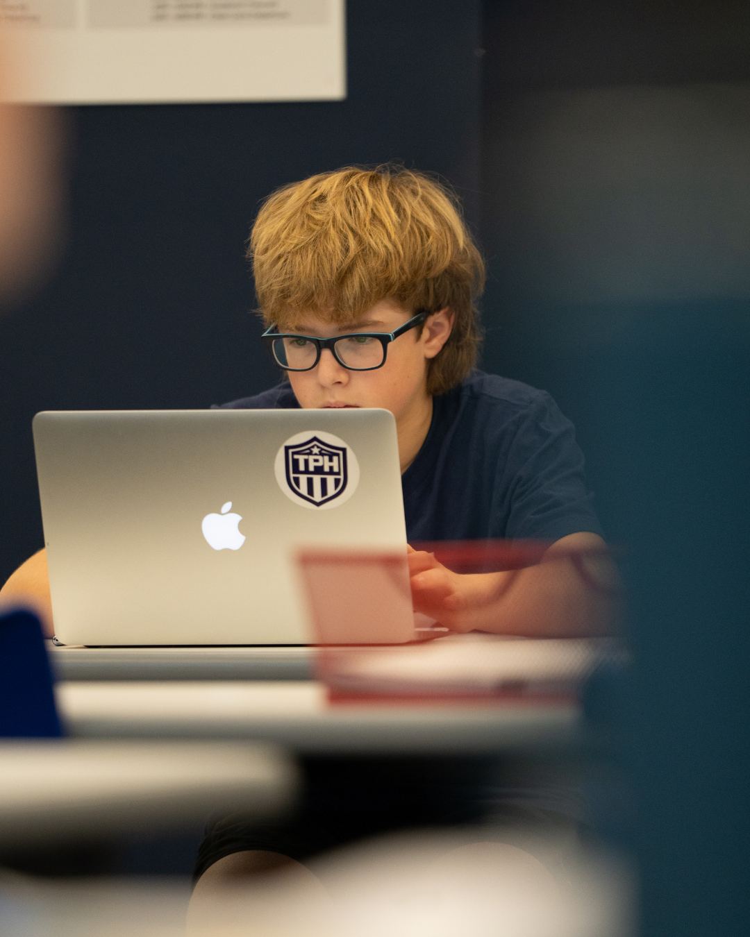 Student with glasses from across the room looking at their open laptop.